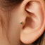 A close-up of a person's ear with a small gold earring featuring a green gemstone in the earlobe.