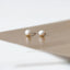 A pair of pearl earrings resting on a gold surface, with the text "SHEMISLI JEWELS" visible below the image.