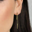 A close-up of a person's ear wearing a pair of gold dangling earrings with a chain and a small pendant.