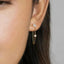 A close-up of a person's ear with a delicate gold earring featuring a small flower-like charm and a dangling chain.