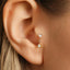 A close-up view of a person's ear, showing a delicate gold earring with two small white stones hanging from it.