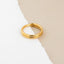 A gold-colored ring is resting on a white surface with a beige background.