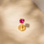 A single, vibrant pink gemstone sits atop a golden, round earring post against a beige background.