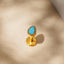 A gold-colored earring with a blue gemstone is shown against a light beige background.