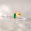 A gold earring with a green gemstone is shown against a blurred, water-like background.