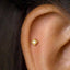 A close-up of a person's ear with a small gold-colored push pin inserted into the skin.
