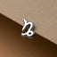 A silver-colored metal charm in the shape of a zodiac sign, specifically Capricorn, is shown against a textured brown background.