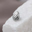 A close-up image of a silver-colored, textured object, possibly a piece of jewelry, resting on a white surface.