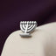 A silver menorah pin is displayed on a white fabric surface against a purple background.