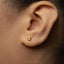 A close-up of a person's ear with a small gold earring featuring a hand-like symbol, likely representing the Hamsie symbol.