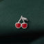 A close-up image of a silver pin with a red cherry design on a dark green fabric background.