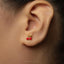 A close-up of a person's ear wearing a small gold earring with a red cherry charm.