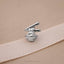 A silver-colored metal pin or badge with a stylized design, resting on a beige fabric surface.