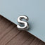 A shiny silver letter 'S' is placed on a textured surface, such as a piece of fabric or paper.
