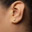 A close-up view of a person's ear, showing a small gold-colored earring with a spade shape.
