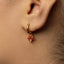 A close-up of a person's ear wearing a gold hoop earring with a red mushroom charm hanging from it.