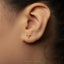 A close-up of a person's ear with a small gold-colored earring featuring a symbol resembling a zodiac sign.