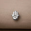 A silver-colored metal pin or badge with a hand-shaped design, featuring an eye symbol, is resting on a beige fabric surface.