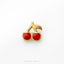 A close-up image of a small, gold-colored pin or brooch in the shape of a pair of cherries.