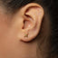 A close-up of a person's ear with a small gold earring featuring a cross-like symbol.