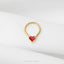 A gold-colored heart-shaped piercing ring with a red gemstone in the center, placed on a white fabric surface.
