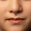 A close-up portrait of a woman with a red gemstone nose ring, looking directly at the camera with a neutral expression.
