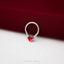 A silver ring with a red heart-shaped charm is placed on a red fabric surface.