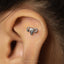a close-up of a person's ear with a small silver earring in the shape of a reproductive organ, specifically a pair of ovaries.