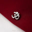 A close-up image of a silver Om symbol pin resting on a red fabric surface.