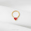 A gold-colored heart-shaped piercing ring with a red gemstone in the center, placed on a white fabric surface.