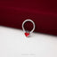 A silver ring with a red heart-shaped charm is placed on a red fabric surface.