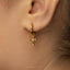 A close-up of a person's ear wearing a gold-colored earring with a small cross-shaped charm hanging from it.