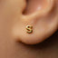 A close-up of a person's ear with a small gold earring featuring the letter 'S' in the center.