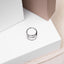 A silver ring is placed on a white surface, surrounded by a beige box and a white background.